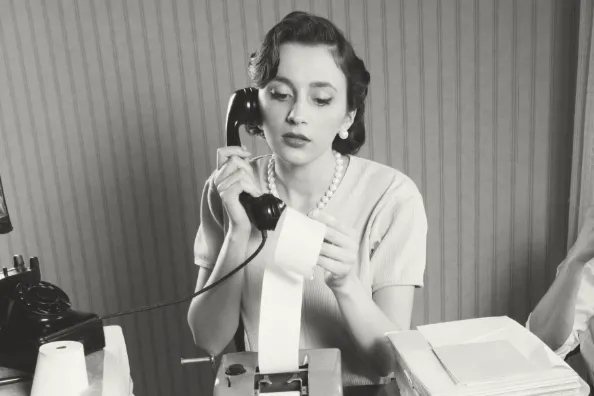 Image d'illustration des coûts de fonctionnement annuel du service Attraction - Jeune femme au téléphone dans les années 50 qui regarde le détail d'un rouleau d'impression d'une calculatrice - Photo noir et blanc
